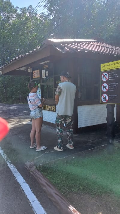 Two people are standing in front of a kiosk or small building in a natural area, likely a park or nature reserve. The person on the right is wearing a light blue shirt, camouflage pants, and a baseball cap. The person on the left is wearing a plaid shirt and denim shorts. The kiosk has signs with regulations in Thai and English. The surrounding environment includes trees and grass, indicating an outdoor setting. The location is likely Ko Panyi, Thailand, based on the Thai language on the signs.