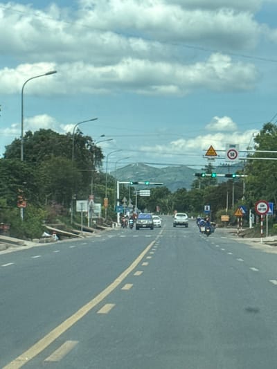The image depicts a straight, asphalt road with a double yellow line in the center and white dashed lines marking the lanes, indicative of a highway or major road. The road is lined with green trees and vegetation on both sides, with some evidence of infrastructure such as streetlights and traffic signals. In the distance, mountains are visible under a partly cloudy sky. Several vehicles, including cars and motorcycles, are present on the road. Traffic signs are also visible, including speed limit signs and what appears to be a "No U-turn" sign. The presence of these elements suggests a typical urban or semi-urban environment in Vietnam. Based on the overall scene, this could be a road within or near Lao Bảo Commune, Quảng Trị Province, Vietnam, known for its roads connecting to the border.