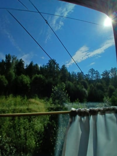 The image captures a view from a window, likely on a train, looking out onto a sunny, green landscape with a bright blue sky. Several taut wires crisscross the sky, suggesting overhead power lines. In the foreground, a thin, light brown rod runs horizontally across the frame, supporting a white, ruffled curtain that partially obscures the lower right portion of the view. Beyond the window, a dense, dark green forest covers a hillside under a clear blue sky dotted with wispy white clouds. The sun is visible in the upper right corner, casting a bright glare and lens flare. The scene is identified as being in Khrebet, Russia.