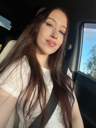 A young woman with long, dark brown hair and fair skin is seen from a selfie perspective inside a car. She is wearing a white t-shirt and has subtle makeup, including winged eyeliner. Her hair is spread across her chest and shoulder. She appears to be looking at the camera with a slight smile.

The scene is illuminated by natural light, suggesting it is daytime. Sunlight is coming from the right, casting shadows and highlighting her features. Through the car's sunroof and possibly window, a bright blue sky with a few wispy clouds is visible, along with the silhouette of tree branches, indicating an outdoor setting. The car's interior is dimly lit in comparison, with a black dashboard and a grey seatbelt partially visible. A beige-colored edge of the car seat is also in the foreground. The overall mood appears relaxed and casual.

There is no visible text in the image.