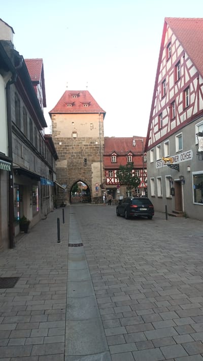 This is a photograph of a street scene in Altdorf bei Nürnberg, Germany. The street is paved with cobblestones and lined with traditional German architecture. On the left, a building with a dark mansard roof and arched windows is visible. To the right of that, a large stone tower with a red tiled roof and dormer windows stands prominently. The tower has a large arched entryway through which more of the street and buildings can be seen. Further to the right, a building with a steeply pitched red tiled roof and white half-timbering is visible. Adjacent to that is a grey building with a red tiled roof and several windows. A black car is parked on the right side of the street. The sky is clear and bright, suggesting it is daytime, likely late afternoon or early evening due to the soft lighting. The scene appears peaceful and quiet, with a few people visible near the arched entryway of the tower. There is text visible on the grey building to the right, which appears to be a sign for "GAS THOF D GNER OCHSE".