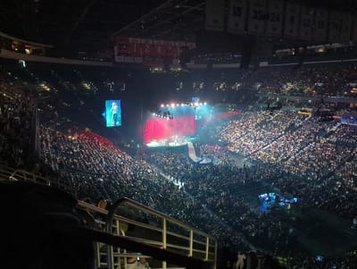 The image displays a large-scale indoor concert event at the Wells Fargo Center in Philadelphia, Pennsylvania, United States. The vantage point is from an elevated seating section, looking down and across the arena bowl, which is filled with thousands of spectators. A primary stage is situated at one end of the arena floor, featuring multiple performers, bright stage lighting, and a prominent red curtain or backdrop. To the left of the stage, a large vertical LED screen displays a close-up image of a female performer with light-colored hair. A catwalk extends from the main stage into the dense crowd on the arena floor. The tiered seating sections surrounding the arena are fully occupied. Numerous small lights, likely from cell phones, are visible throughout the audience. Banners displaying retired numbers (e.g., "15", "20", "24") associated with the Philadelphia Flyers hockey team are visible near the arena ceiling. A metal railing from the viewing section is present in the foreground.
