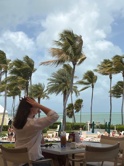A horizontal, eye-level shot captures a scene at the beach in Key Biscayne, Florida. Palm trees line the shore, with the ocean visible in the background, alongside sunbathers in lounge chairs. A woman is visible, seated at a table, with her hand on her head. The table also contains items like a menu and sunglasses.
