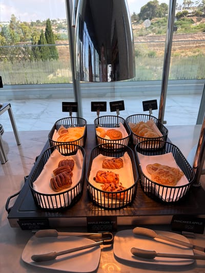 This is a close-up, eye-level, indoor shot of a buffet display of baked goods, likely in a hotel or restaurant. The baked goods are arranged in black wire baskets on a heated buffet serving station. From left to right, the visible labels indicate "PANCAKE CLASSIC", "OLIVE PIE", "TAHINI PIE", "SPINACH & CHEESE PIE", "APPLE PIE", and "PANCAKES APPLE".

In the foreground, two pairs of serving tongs rest on white plates, ready for use. The backdrop consists of a large window showcasing a landscape of green trees and a distant urban area with buildings, possibly the city of Ayia Napa, Cyprus, under a bright, daytime sky. A tall, metallic structure, part of the buffet's lighting or support system, is prominently reflected in the window. The overall scene suggests a self-service breakfast or brunch offering. No people are visible in the frame.