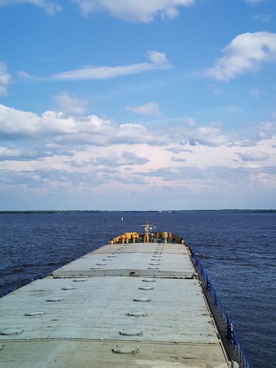 This is a photo taken from the deck of a barge traveling on the Volga River near the village of Veregonets, Russia. The sky is blue with scattered white clouds. The river is dark blue and choppy. In the distance, there is a green shoreline. The barge is grey and has a yellow railing and several circular hatches on its deck. There are no people visible in the image.