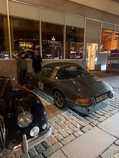 The image captures an evening scene in Sankt Moritz, Switzerland, on Via Maistra 25, in front of the Crystal Piano Bar, part of the Crystal Hotel.

In the foreground on the left, the front section of a dark-colored classic automobile is visible. It features prominent chrome-ringed headlights, a chrome bumper, and a partial view of its grille. A white rectangular plate with a red number "1" is partially observable below the bumper.

In the midground, a dark grey Porsche 911 Targa is parked, angled with its rear towards the viewer. The car displays a white circular "43" on its driver's side door and an orange "43" sticker on its rear window, indicating participation in an event. It bears a Swiss license plate, "ZG-5047." The vehicle has a black Targa roof panel, a louvered engine cover, and chrome wheels with black centers.

Two adult males are standing on the paved sidewalk between the building facade and the Porsche. The man on the left wears a dark beanie and a dark green jacket, standing with his hands in his pockets. The man on the right wears a light grey beanie with ear flaps, a dark jacket, and an orange neck garment, and is gesturing with his right hand towards the Porsche while facing the other man.

Behind the individuals, the Crystal Piano Bar is visible through large, rectangular glass windows. The interior is illuminated, revealing a well-stocked bar with numerous liquor bottles. A lighted sign displaying "Crystal Piano Bar" and three stars is prominent. To the right of the bar windows, a silver-framed glass door is marked with "TAVOLATA" and a stylized sun logo. Further right, a wooden display stand with partially visible text, possibly "#RentLike", is situated on the sidewalk. The ground surface consists of light-colored cobblestones in the street and a smoother, lighter paved section for the sidewalk adjacent to the building.