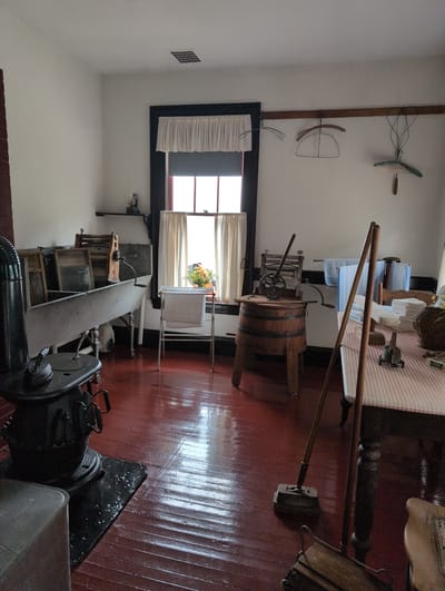This image depicts a room that appears to be a historical laundry or washroom, possibly located in Campobello Parish, Canada. The room has white walls and a rich, glossy red wooden floor that reflects the light.
Dominating the left side of the image is a large, old-fashioned wash tub with a metal basin and white pedestal legs. Next to it, to the left, is a black cast-iron stove with a chimney pipe extending upwards. Several washboards are propped inside or against the wash tub.
In the center of the room, a large window with a dark frame is covered by a white valance at the top and cream-colored curtains. A small vase with yellow and orange flowers sits on the windowsill. To the right of the window, a chrome drying rack holds a white towel.
Further to the right, a wooden barrel serves as a stand for a vintage washing machine with a hand crank and rollers. Next to this barrel, a wooden table is covered with a red and white checkered cloth. On the table, there are stacks of folded white cloths, a basket, and various laundry-related items, including what appear to be flat irons.
Two long wooden-handled brooms with brush heads stand upright on the floor in the foreground, leaning against the table. Behind these, a piece of furniture with a woven seat is visible.
Hanging from a wooden rod on the wall above the window are several antique wire and wooden clothes hangers. The overall impression is of a preserved historical domestic space, likely a museum exhibit or a carefully maintained heritage home. There are no people visible in the scene.