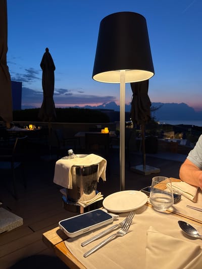 This is a photograph taken outdoors at dusk, likely from a restaurant terrace in Crveni Vrh, Croatia, overlooking the sea. The foreground features a table set for a meal. There is a white plate, a glass of water with condensation, a fork, and a spoon on a beige placemat. A mobile phone lies on the table. To the left, an ice bucket contains a bottle of what appears to be sparkling wine, covered with a white cloth. In the background, there are outdoor umbrellas, tables, chairs, and a large floor lamp with a black shade. The sky is a deep blue transitioning to shades of purple and orange near the horizon, with silhouetted clouds. The sea and a distant coastline are visible. A person's arm and shoulder, wearing a light blue polo shirt, are visible on the right side of the frame, suggesting they are seated at the table.
