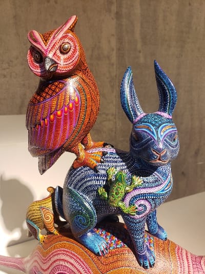 The image shows ornately decorated wooden animal sculptures in vibrant colors. A blue rabbit-like figure with long ears balances a green frog on its back and hangs onto a larger, abstract creature. Below, an orange owl-like figure faces left. The sculptures are covered in intricate patterns and details. The background is a neutral gray wall.