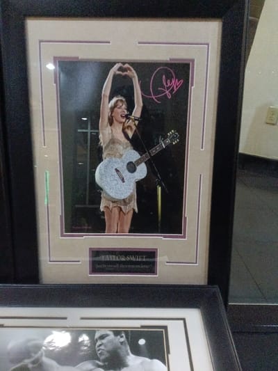 A framed and autographed photo of Taylor Swift performing with a guitar and making a heart shape with her hands is on top. The frame has multiple layers of beige and purple matting. A plaque below the image reads "Taylor Swift, Just be yourself, there is no one better." Underneath the Taylor Swift framed picture, a second black frame is partially visible showing a black and white photo of Muhammad Ali in a boxing stance. A wall with an electrical outlet is partially visible in the background.