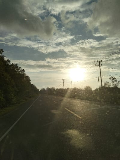 The image captures a rural road scene in Keudeunga, Indonesia, viewed from what appears to be a moving vehicle. The primary subject is a two-lane asphalt road extending into the distance, marked by a continuous white line on the left shoulder and intermittent yellow dashed lines indicating the center. The road surface appears wet or highly reflective, possibly due to recent rain or dew, with sunlight reflecting prominently. The sun is low in the sky, positioned toward the upper right, generating significant lens flare and a bright glare that partially obscures the view.

To the left of the road, a densely vegetated hillside covered in trees and bushes rises. To the right, multiple wooden utility poles supporting numerous power lines run parallel to the road, with additional green foliage and smaller trees visible behind them. The sky above is partially cloudy, featuring a mix of darker, heavier clouds and lighter, scattered cloud formations, indicating either sunrise or sunset conditions. No individuals or other vehicles are visible on the road.