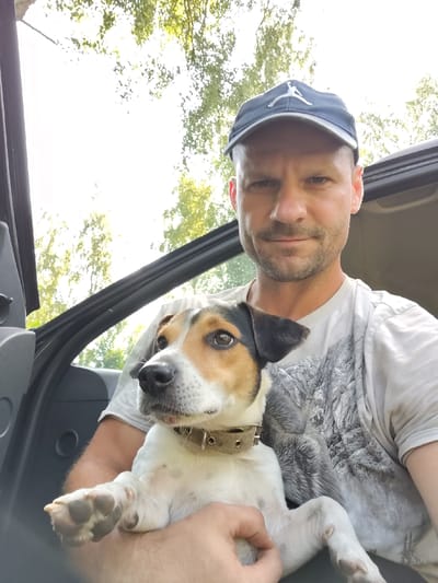 A middle-aged Caucasian male with short, light brown hair, stubble, and a light beard is seated in the driver's seat of a vehicle. He is wearing a grey t-shirt with a grey abstract design and a dark blue baseball cap with a white Jumpman logo. He is holding a small terrier mix dog with white, tan, and black markings on its face and body. The dog is wearing a tan collar and is looking towards the camera. The interior of the vehicle, including the door panel, is visible. The background shows bright sunlight filtering through green foliage, suggesting an outdoor setting during the day. The specific location in Чарацянскі сельскі Савет, Belarus, cannot be determined from the visual information provided in the image.