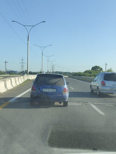 The image depicts a daytime scene on a highway, likely in or near Sannikovo, Russia. The sky is clear blue, and the sun is visible, casting shadows. Several streetlights with multiple arms line the side of the road. In the foreground, on the right side of the frame, a silver compact SUV is visible driving on the highway. To the left, a blue hatchback is also driving on the highway. The hatchback has a distinctive dark pattern applied to its rear bumper and a visible license plate that reads "M718 TK 54". The road itself is asphalt, marked with white lines. In the background, there are power lines, utility poles, and trees. The overall impression is of a typical highway environment under clear weather conditions.