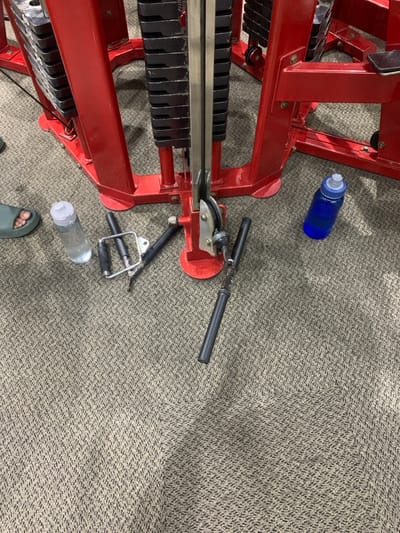 The image shows a red weight machine in a gym in Kecamatan Kartasura, Indonesia (latitude: -7.567, longitude: 110.757). The machine has a cable system and weight stacks on either side. Two black handles are lying on the floor next to the machine, and a blue water bottle is sitting on the carpeted floor. A clear plastic water bottle is laying on the floor in the left side of the image. A bare foot and a blue flip flop can be seen in the bottom left corner. The scene is a gym, and the image is taken from a low angle. The floor is carpeted with a light brown and black pattern.