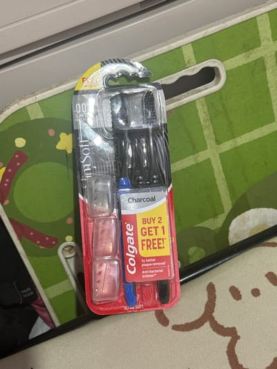 An overhead medium shot shows a Colgate toothbrush multipack, with a "Buy 2 Get 1 Free" sticker, lying on a green and yellow checkered surface with brown abstract patterns. The toothbrush is in transparent packaging and the package is red and yellow. The toothbrush has a black handle and a blue bristle head, with two additional toothbrush covers. The background is a blurred, gray surface. The overall scene appears to be an indoor setting, possibly a table or counter, in a city like Manila, Philippines, indicated by the common availability of such products and the general domestic environment.