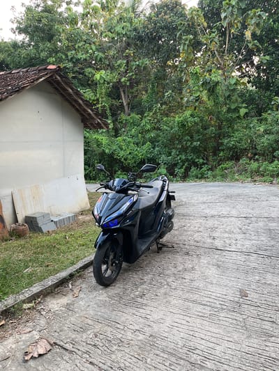 In the city of Legonipah, Indonesia, a black scooter with blue accents on its headlights is parked on a rough concrete surface. To the left of the scooter is a small, light-colored building with a terracotta tile roof. Next to the building, on the grassy ground, are several stacks of grey tiles and a terracotta pot. The background is filled with dense green trees and foliage, suggesting a natural, possibly rural or semi-rural setting. The sky is overcast.