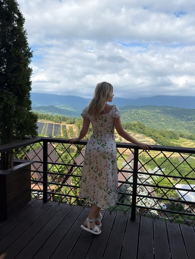 A woman with blonde hair, wearing a white floral dress and white sandals, stands on a wooden balcony overlooking a valley. The balcony has a dark wood floor and a black metal railing with a diamond pattern. In the background, rolling green hills are covered in trees and some fields. A large evergreen tree stands to the left of the balcony. The sky is partly cloudy. The location is identified as Nizhnyaya Shilovka, Russia.