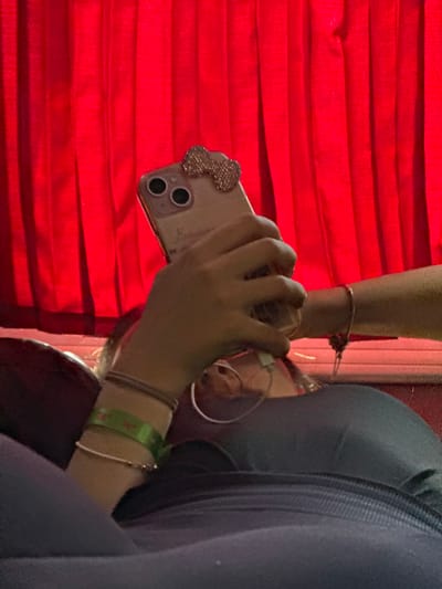 A person is holding an iPhone in front of a red, horizontally-striped surface. The phone has a jeweled decoration in the upper left corner and dual cameras. The person is wearing multiple bracelets on their left wrist, including a green wristband with a red design. The setting appears to be indoors, in the city of Izvorovo, Bulgaria.