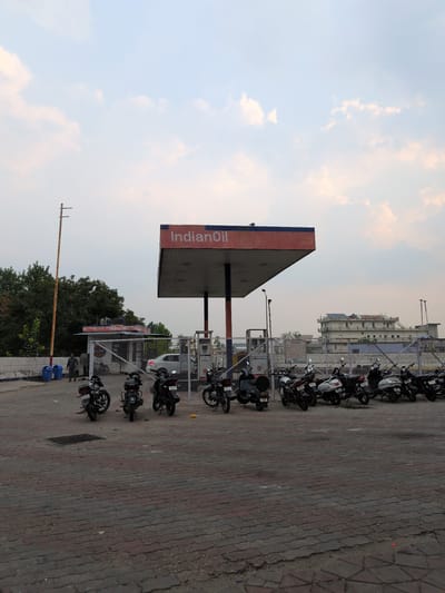 An exterior, eye-level, vertical shot captures an IndianOil gas station under a partly cloudy sky in Una, India. The gas station canopy, with its prominent "IndianOil" logo, is the central focus. Several motorcycles are parked in rows in front of a chain-link fence. To the left, a utility pole stands tall. In the background, a multi-story building, possibly a hospital, is visible. The ground is paved with interlocking bricks. The scene is likely captured during daylight hours, given the illumination.