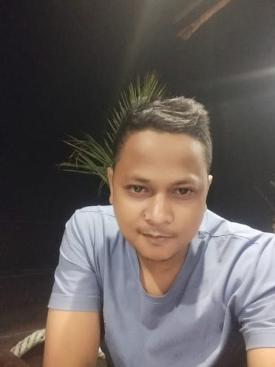 A close-up, dimly lit portrait features an adult man, likely of Southeast Asian descent, looking directly at the camera. He has short, dark hair, dark eyes, a faint mustache, and light stubble, with clear skin. He wears a light blue, short-sleeved t-shirt. The overall lighting suggests the photo was taken at night or in a very dark environment, with direct light (possibly a camera flash) illuminating his face and upper body.
Behind and to the left of his head, discernible green fronds, characteristic of palm trees, are visible against the dark background. To the right, above his head, there's a faint indication of a linear wooden structure. Below his right arm, a thick, braided white rope is visible, suggesting a setting near water, such as a dock, boat, or coastal area.
The man maintains a neutral expression, with perhaps a very slight, subtle hint of a smile. The context of Keudekruenggeukueh, Indonesia, supports a tropical, coastal location for this nighttime scene. There is no visible text in the image.