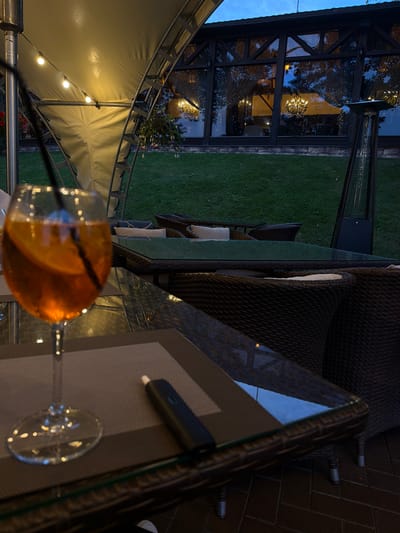 The image depicts an outdoor seating area at dusk, likely a restaurant or cafe, in Izmailovo, Russia. The scene is set on a patio with a brick floor and a grassy lawn in the background. A glass-topped table is in the foreground, with a woven chair and cushions visible. A cocktail with an orange slice sits in a stemmed glass on the table, along with what appears to be a vaping device. String lights are visible under a large, white tent-like structure, suggesting an evening ambiance. Behind the table, a modern glass-enclosed structure with interior lighting and a chandelier is visible, and beyond that, a well-maintained lawn. The lighting indicates it is twilight, with the sky transitioning from blue to a darker hue. The overall impression is one of a relaxed evening outdoor dining or lounging experience. There is no visible text in the image.