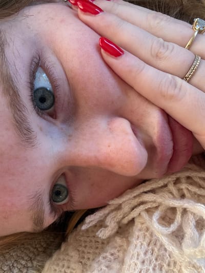 A close-up, oblique photograph captures a person's partially obscured face. The scene is reportedly located in Rosmalen, Netherlands; however, the image itself lacks specific environmental indicators for this location. The subject's right hand, displaying long, oval-shaped fingernails painted a glossy red, is placed across the upper portion of the face, covering the forehead and temple area. Two open blue-grey eyes with visible eyelashes are directed upwards. Fair skin with minor moles or freckles is discernible on the nose and cheek. The bridge and tip of the nose are visible, along with a portion of the subject's pinkish lips. On the right ring finger, two gold-toned rings are worn: one is a narrow, textured band, and the other features a prominent, light-colored, possibly clear, faceted stone set in a raised mounting. Reddish-brown hair is visible along the lower-left edge and beneath the hand. A cream-colored, fringed knit garment is present in the lower right quadrant of the image, appearing to rest against the subject's neck or shoulder.