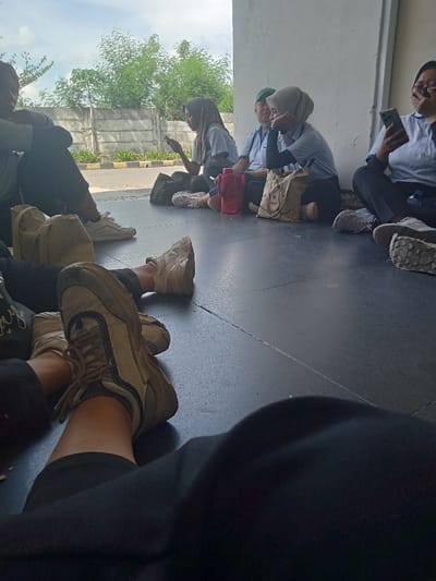 This image depicts six individuals seated on a dark, tiled floor under a covered structure, likely in Sukomoro, Indonesia, with a seventh person's legs visible in the immediate foreground. The perspective is from a low vantage point, looking outwards towards a bright, sunny exterior.
From left to right:
1. **Left foreground:** The lower legs and feet of an individual wearing black pants and beige-and-dark-accented sneakers are stretched out towards the bottom-center of the frame.
2. **Far left:** A person, largely obscured, is visible from the waist down, wearing dark pants and white sneakers, seated on the floor.
3. **Left-center:** A female, wearing a light blue short-sleeved polo shirt, dark pants, and a light grey hijab, is seated cross-legged on the floor. She is looking down at a smartphone held in both hands. A dark bag rests beside her left leg.
4. **Mid-center:** A male, wearing a light blue short-sleeved polo shirt and a green cap, is seated on the floor with his right arm resting on his bent knee, looking towards the left.
5. **Right-center:** A female, wearing a light blue short-sleeved polo shirt over a dark long-sleeved undershirt, and a light grey hijab, is seated on the floor with her chin resting on her left hand, looking towards the left. A red thermal flask and a light brown fabric tote bag with a graphic design are positioned next to her right leg.
6. **Far right:** A female, wearing a light blue short-sleeved polo shirt and dark pants, is seated on the floor, possibly cross-legged, holding a smartphone that partially covers her face as she looks downwards. A blue plastic bottle is on the floor near her right leg.
The individuals appear to be resting or waiting. The background reveals an outdoor scene with an asphalt or paved surface, a low concrete wall or fence, and dense green trees and foliage under a bright, cloudy sky.