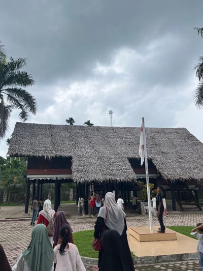 A group of people, many wearing hijabs, walk on a stone pathway towards a traditional Indonesian house with a thatched roof. A flag flies from a pole in front of the house. In the background, palm trees and other greenery are visible under a cloudy sky. A cell tower is also visible in the distance. The scene appears to be an outdoor cultural site or historical landmark.