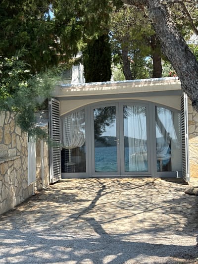 This is an eye-level, medium shot of a modern building with large glass doors facing a body of water, likely the Adriatic Sea, in Grad Šibenik, Croatia. The building's exterior is made of light-colored stone, and it is partially obscured by lush green trees. The foreground is a stone patio with a gravel border, and tree shadows are cast across the patio. The glass doors are framed by gray shutters, one of which is open. Inside the building, sheer white curtains are visible. The scene appears to be a residential or commercial property, possibly a vacation rental, given its waterfront location and modern design.
