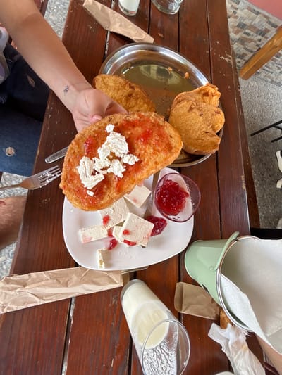 An overhead view depicts a rustic meal laid out on a dark brown slatted wooden table in Zasele, Bulgaria. A person's left arm and hand, adorned with a silver bracelet featuring a small charm, are visible on the left, holding a large piece of golden-brown fried bread topped with red jam and crumbled white cheese. Dark blue ripped jeans are visible on the person's lap. To the immediate left, a silver fork and knife rest on the table, the fork showing white residue. Further left, a male's hairy right leg is partially visible under the table.
On the table, a white rectangular plate holds multiple blocks of white cheese, some speckled with red jam. Adjacent to this plate, a small clear glass bowl contains a portion of red jam. In the upper center, a round silver metallic tray holds additional pieces of golden-brown fried bread. A tall, clear glass, approximately half-full with a white opaque liquid (likely milk), is situated in the lower left quadrant. Another clear, mostly empty glass is partially visible in the upper left.
The lower right side of the table features a light green enameled metal pail containing white paper napkins, functioning as a napkin holder. Several crumpled brown paper items, possibly wrappers or discarded napkins, are scattered across the table, along with one partially used white paper napkin near the green pail. The floor beneath the table is composed of light and dark gray mosaic-patterned tiles. A dark metal chair leg is visible on the right, and a wooden chair leg is partially visible in the upper right corner.