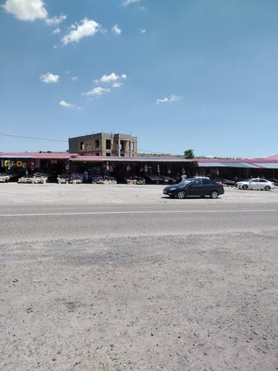 A bright, sunny day is depicted in this image, set in Amankutan, Uzbekistan. The scene is a roadside market with several stalls lined up, showcasing a variety of goods. Behind the market, a multi-story building under construction is visible. The sky is a vibrant blue, dotted with fluffy white clouds, suggesting fair weather.
In the foreground, a paved road with a white line runs horizontally across the frame. Beyond the road lies a gravel area where the market stalls are situated. The stalls themselves are covered by colorful awnings in shades of red, green, and white. They display numerous items, including what appear to be sacks of produce, bags of goods, and brightly colored inflatable rings. A few people are seen near the stalls, some standing and interacting with vendors.
Two cars are present in the image. A dark-colored sedan is parked in front of the market stalls, and a white sedan is further down the road. The overall atmosphere seems to be that of a typical day at a bustling roadside market. The construction of the building behind the market hints at ongoing development in the area. The time of day is clearly daytime, indicated by the bright sunlight and clear sky.