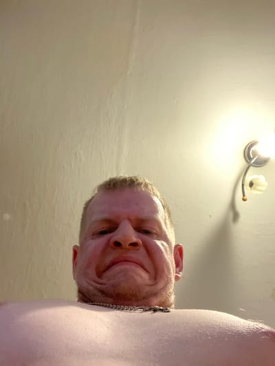 A low-angle, close-up shot shows the chest and lower face of a fair-skinned man with short, light brown hair. He is wearing a silver chain necklace. His face is contorted into a grimace, with his mouth pulled down at the corners and his cheeks bunched up. The background is a textured, cream-colored wall with a light fixture on the right side. The lighting is warm and appears to be from an artificial source. The location is Unknown, Unknown.