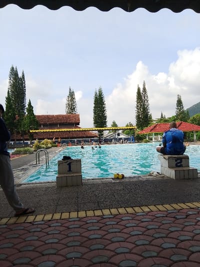 This image depicts an outdoor swimming pool complex located in Ciputri, Indonesia, viewed from beneath a dark, wavy-edged overhang. In the immediate foreground, a patterned pavement features red and gray fan-shaped tiles with a yellow border. A partially obscured person in gray pants and dark flip-flops stands on the left. Centrally, two white concrete swimming starting blocks are visible, labeled '1' and '2'. A person in a blue shirt with a white graphic on the back is seated on block '2', facing away from the viewer. A yellow floating object and white flip-flops rest on the pool deck near block '1'. The rectangular pool contains light blue water with several individuals swimming or standing. Beyond the pool, a building with a brown-tiled roof and a yellow extended awning is present, flanked by tall coniferous trees. Further structures, including a white building with a blue roof and a red-tiled gazebo, are visible. The background reveals a partly cloudy sky and a green, tree-covered mountain on the right.