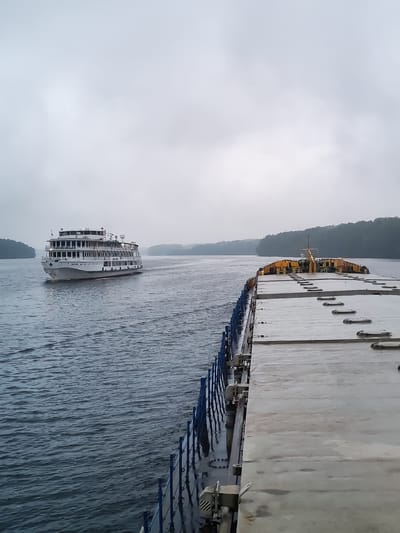 In the city of Ankimarovo, Russia, on a body of water under an overcast sky, two vessels are visible. On the left, a large, white multi-decked passenger ship sails away from the viewer, its multiple windows and upper decks clearly defined. On the right, a portion of a different vessel, likely a cargo barge, is in the foreground, its flat, grey deck featuring numerous latches and hatches. The barge's blue railing runs along its side. The water around both vessels appears choppy, indicating movement. The distant shoreline is lined with dense green trees.