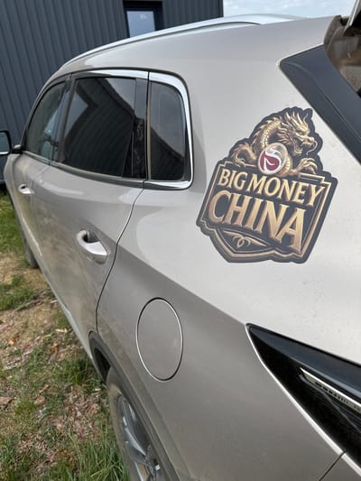 In Khuzhir, Russia, a silver SUV is parked outdoors on a grassy area. The vehicle has a sticker on its rear passenger door that reads "BIG MONEY CHINA" in gold lettering, featuring a golden dragon design and a red and white circular emblem. The SUV is positioned with its rear quarter visible, and the tailgate is slightly ajar. The background shows a dark-colored building with a window. The overall scene suggests an exterior setting, possibly a residential or private property in Khuzhir.