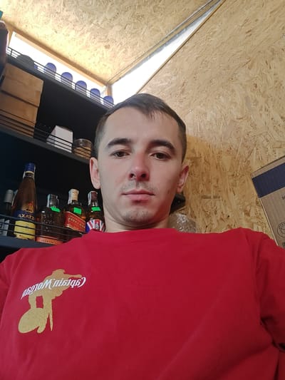 A man is taking a selfie in front of shelves stocked with bottles and boxes. The shelves are mounted on a wall made of particleboard. The man is wearing a red shirt with a gold graphic of a person on a surfboard. The background includes what appears to be a window at the top, letting in natural light. The scene is likely indoors, possibly a pantry or storage area.