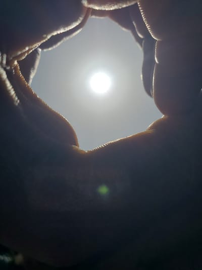 The image shows a close-up view of a person's hands forming a circular shape, framing a bright sun against a hazy sky. The sun is the primary light source, creating lens flare and highlighting the edges of the fingers with a bright, almost sparkling effect. The hands are in silhouette, with their textures and contours visible due to the backlighting. The overall impression is one of looking through a hand-made frame at the sun. The context suggests this could be a moment of observation or contemplation, possibly during the day. The haziness of the sky could indicate a particular weather condition or time of day. There is no visible text in the image.