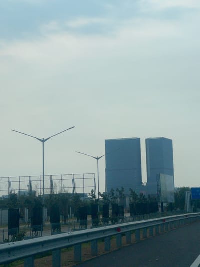 The image captures a highway scene in Kosambi Barat, Indonesia, with two large, dark blue rectangular buildings dominating the background. These buildings appear to be skyscrapers, possibly office towers or residential complexes, and are situated on the right side of the frame. In the mid-ground, a series of streetlights with multiple arms are visible, some extending over the highway. A metal guardrail runs along the edge of the road in the foreground, separating it from a grassy verge and a fence. Behind the fence, there are dark, rectangular structures interspersed with trees and foliage. The sky is overcast with pale clouds, suggesting a muted or hazy day. There are no people or vehicles clearly visible in the scene, and the overall atmosphere is one of urban infrastructure and development. A blue sign is visible on the far right with text that is partially legible.