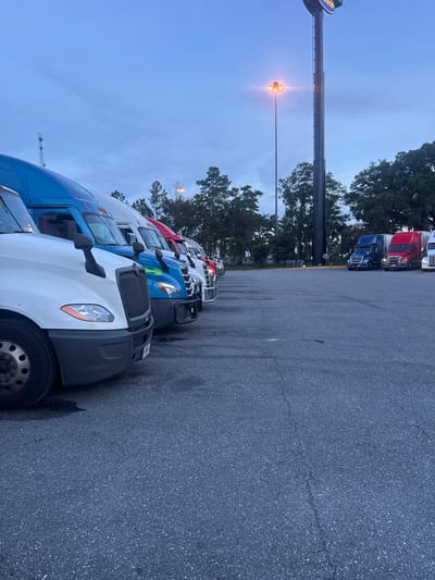 A row of semi-trucks are parked in an asphalt lot at dusk. The trucks are facing the viewer from right to left, with the closest truck on the left being white. Behind the white truck are blue, green, and red trucks. In the background, trees and a large tower with a sign are visible. A tall pole with lights stands in the middle ground. The sky is a pale blue with some clouds. The scene appears to be at a truck stop or rest area. The sign on the tower appears to say "LOVE'S" which is a chain of travel stops located throughout the United States, including in Lake City, Florida.