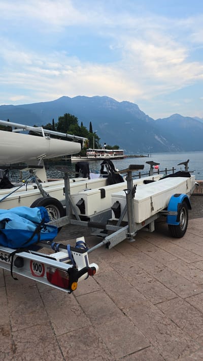 This media file shows a trailer with a sailboat on it, parked on a stone-paved area next to a body of water. In the background, there are mountains and a cloudy sky. A boat is docked in the water. The trailer is white and metallic gray, with blue accents and a red taillight. A "100" sticker is visible on the trailer. The scene appears to be set at Riva del Garda, Italy. The time of day is likely late afternoon or early evening, given the soft lighting and the color of the sky. The weather seems to be mild, with some clouds present. No people are visible in the image, but the presence of the trailer and sailboat suggests an activity related to boating or water sports. The overall mood is peaceful and serene, with a focus on the natural landscape and the equipment for recreation.