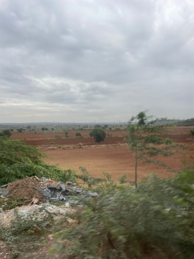 The image depicts a wide, rural landscape under a cloudy sky. The foreground is dominated by a pile of debris, including broken concrete, bricks, and what appears to be discarded plastic sheeting, partially obscured by green foliage. Behind the debris, a field of dry, golden-brown grass stretches into the distance. Further back, large areas of reddish-brown soil, possibly cultivated fields, are visible, interspersed with scattered green trees. In the far distance, a hazy horizon line suggests a populated area with some structures and possibly power lines or industrial elements. The overall impression is of an undeveloped or semi-developed rural environment on the outskirts of Bethamcherla, India, characterized by agricultural land, natural vegetation, and accumulated waste.