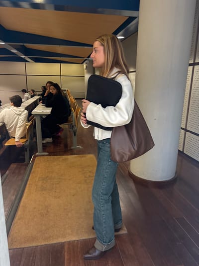 A young woman stands in profile on the right side of a lecture hall in Porto Salvo, Portugal, facing left. She has long, light brown hair and is dressed in a white collared jacket, blue jeans with cuffed hems, and dark brown pointed-toe boots. She holds a black rectangular folder or laptop case in her left arm and carries a large, dark brown, textured leather tote bag on her left shoulder. To her left, a long, light brown mat rests on dark wooden floorboards, defining an aisle. Beyond the aisle, multiple students are seated at tiered rows of light wooden desks with metal frames. One male student in a white hoodie is visible from the back, while a female student with dark hair, wearing a dark top, is seated further back, looking directly towards the camera's general direction. Other students are partially visible in the background. A large, light-colored cylindrical pillar is positioned to the right of the standing woman. The room's ceiling features blue and light wood-toned panels with recessed lighting, and the back wall exhibits a perforated or grid-like pattern in a light color.