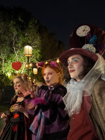 This outdoor night image, captured in Nir Eliyahu, Israel, displays three individuals dressed in elaborate costumes, strongly suggesting an "Alice in Wonderland" theme. They are posing for a photograph in what appears to be a festive, illuminated garden setting.
From left to right (when the image is viewed upright):
The first person is costumed as the Mad Hatter. They wear a reddish-brown top hat adorned with a white clock face, showing hands pointing to approximately 7:00. The hat also features blue and purple feathers and a fluffy grey pom-pom. This individual has pale skin, light blue eyeshadow, and reddish lipstick, and is wearing a reddish-brown jacket with a sheer white ruff.
The middle person is dressed as the Cheshire Cat. They have reddish hair with purple ear-like accessories and prominent cat-like facial makeup, including drawn whiskers and a wide, toothy grin. They are wearing a purple and black striped garment.
The third person on the right is costumed as the Queen of Hearts. They have blonde hair styled up, topped with a small gold crown and a red glittery heart on a headband. Their face is decorated with small red hearts on the cheeks and bold red lipstick. They wear a black dress embellished with red heart designs and are holding a decorative golden key or scepter.
The background is dark, indicating night, with warm string lights and rectangular lantern-style lights hanging amongst lush green trees and foliage, illuminating the scene. The overall mood is whimsical and celebratory, likely from a themed event or party.
