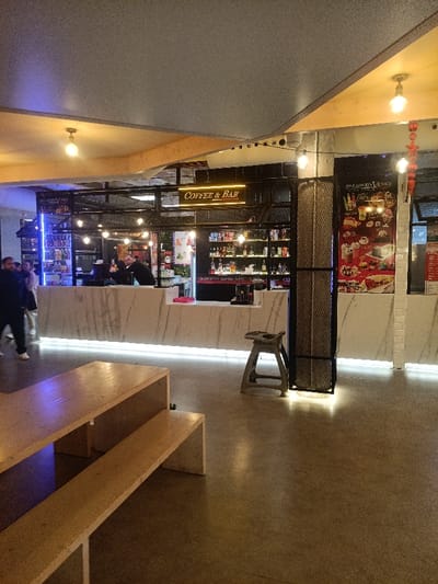 A coffee and bar area showcases a well-lit marble countertop and shelves stocked with beverages. An industrial-style stool sits nearby. A wooden table with an attached bench occupies the foreground. The area is well-lit with both natural and artificial light, casting reflections on the polished floor. A large advertisement for "Gelateria Venice" is visible on the wall.