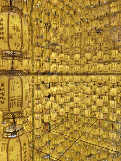 The image displays a dazzling display of numerous yellow lanterns, casting a warm glow. These lanterns are densely packed and appear to be suspended from above, creating a visually stunning effect. The lanterns are cylindrical with decorative elements at the top and bottom, and some feature Chinese characters. The reflections of the lanterns on a polished surface below create an illusion of an even larger, almost infinite collection. The background is filled with more lanterns, creating a sense of depth and immersion. There are no people visible in the image. The lighting is warm and predominantly yellow, emanating from the lanterns themselves. The overall impression is one of a festive or celebratory atmosphere, possibly in an indoor setting such as a market, temple, or exhibition space. The scene is located in Gongshu District, China.