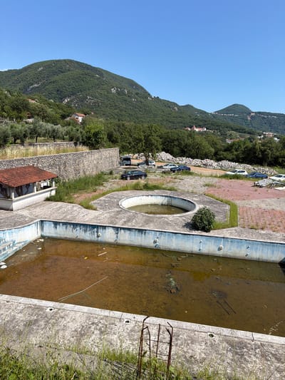 The image shows an abandoned outdoor swimming pool set against a backdrop of lush green mountains and a clear blue sky. The pool itself is empty, filled with murky, stagnant water, and debris. A ladder is visible submerged in the water. The surrounding area is overgrown with weeds and grass, and a stone wall runs along the edge of the property. A few parked cars are visible on a paved area behind the pool. In the distance, houses are scattered on the mountainside, with olive trees growing in the foreground. The scene appears to be in a rural, mountainous area, possibly a resort that has fallen into disrepair. The overall impression is one of neglect and abandonment. The time of day appears to be daytime, with bright sunlight suggesting good weather. There is no visible text in the image.