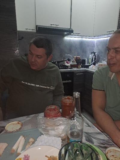 Two men are sitting at a table laden with food and condiments. A bottle of alcohol lies on its side near jars of what appears to be homemade salsa. Plates with food remnants are scattered across the table's surface. The setting is a kitchen, indicated by the presence of cabinets and countertops in the background. The image is slightly tilted.