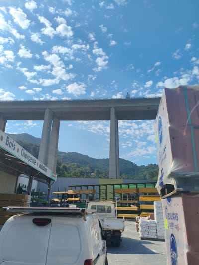 The image depicts a construction supply yard in La Trinité, France, with a large elevated highway overpass dominating the background. In the foreground, a white van is parked, facing away from the camera, with a rack on its roof. To the right, stacks of building materials, including what appear to be bricks and insulation bags, are wrapped in plastic. In the middle ground, several trucks are present, including a white flatbed truck with materials on its bed. Shelves stocked with lumber and other construction supplies are visible behind the trucks. The scene is set against a backdrop of a lush, green hillside under a bright blue sky with scattered white clouds. The signage on the building to the left indicates "Bois Charpente Isolation," suggesting it is a lumber, carpentry, and insulation supply store.