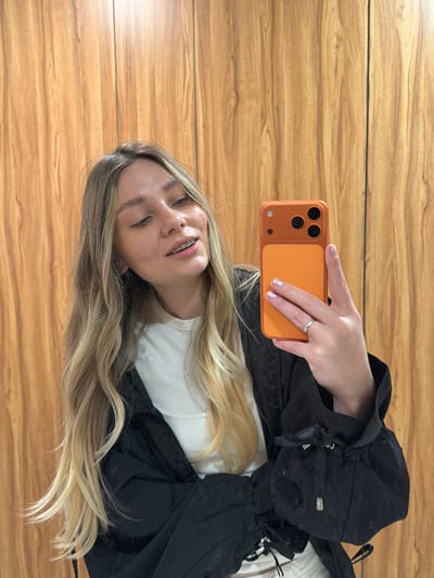 A young adult female is depicted in an indoor setting, taking a selfie with an orange-cased smartphone. She has long, wavy blonde hair and is wearing a white ribbed tank top under a black jacket with ruched sleeves and silver toggles. Visible dental braces are present on her upper teeth. Her right hand, adorned with light nail polish and a silver-colored ring on the ring finger, holds the smartphone. The phone case features a two-tone orange design with three prominent black camera lenses. The background consists of a vertical wooden paneled wall with a medium brown tone and natural grain. The location is Ness Ziona, Israel.
