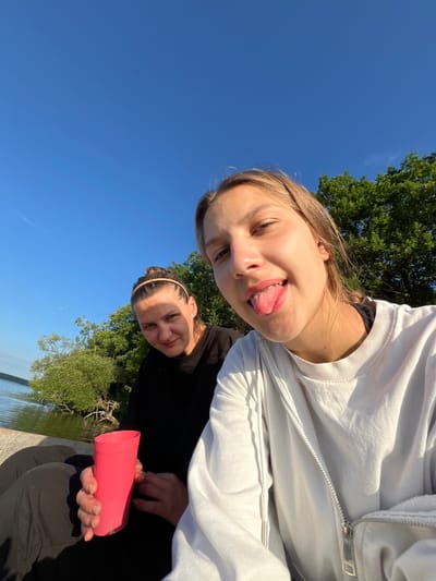 Two young women are seated on a wooden dock by a body of water, likely a lake or river. The woman on the right is closer to the camera and sticking her tongue out playfully, while the woman on the left looks towards the camera with a slight smile. The woman on the left is holding a pink plastic cup. Both are dressed casually in dark and light colored sweatshirts. The background features lush green trees and a clear blue sky, suggesting a warm, sunny day. The water appears calm, reflecting the sky and surrounding foliage. The location is identified as Вяззеўскі сельскі Савет, Belarus, implying an outdoor recreational setting.
